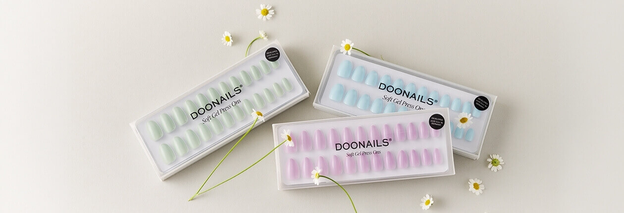 Drei Packungen Doonails Soft Gel Press-On Nails in Salbeigrün, Hellblau und Rosa, dekoriert mit kleinen Gänseblümchen auf hellem Untergrund.