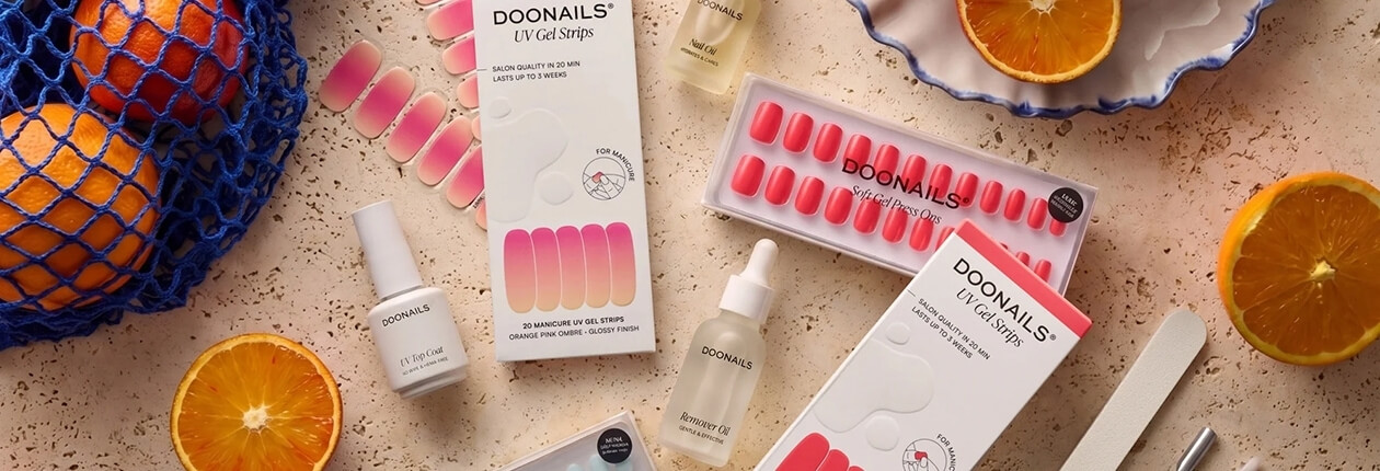 Draufsicht auf Doonails UV-Gel-Strips, Nagellack-Fläschchen, Nagelöl und Orangenhälften auf hellem Untergrund.