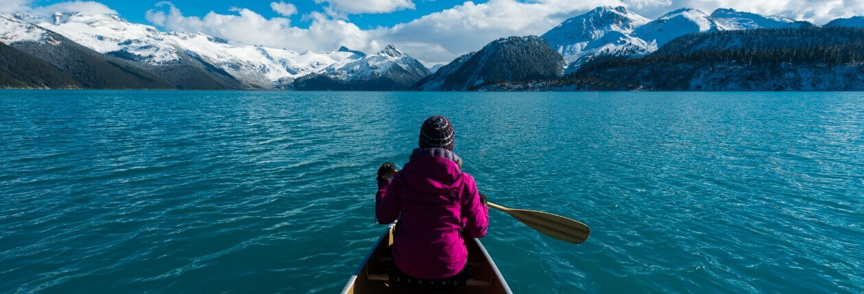 Person in pinker Jacke paddelt in einem Kanu auf einem türkisblauen See vor verschneiter Bergkulisse.